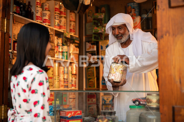 ذكرى تأسيس الدولة السعودية الأولى في 22 فبراير, الأسواق والأحياء التراثية في المملكة, ارتداء الأزياء التقليدية في الأعياد والمناسبات الوطنية, صورة مقربة من الجانب لفتاة عربية خليجية سعودية ترتدي الزي التقليدي تقوم بشراء الحلوى من الدكان الشعبي, يوم بدينا 1727م