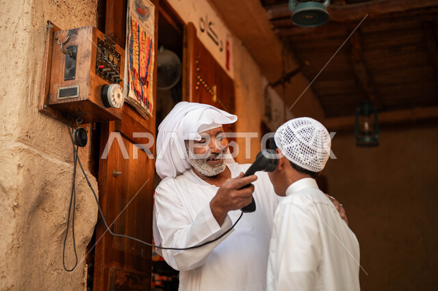 يوم بدينا 1727م, التحدث والتكلم عبر الهاتف الأرضي في الأسواق التراثية, صورة مقربة لرجل كبير بالسن عربي خليجي سعودي يرتدي الزي التقليدي يقوم باستخدام وسائل الاتصال القديمة برفقة حفيده, شعبيات وأدوات نادرة, ذكرى تأسيس الدولة السعودية الأولى في 22 فبراير