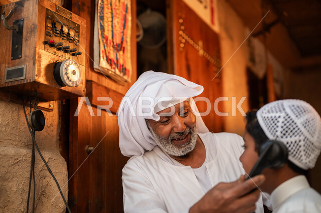 Talking and talking on the landline, a close-up of an old Saudi Gulf Arab man wearing traditional clothing, using old means of communication accompanied by his grandson, folklore and rare tools, the anniversary of the founding of the first Saudi state on February 22, Day of Dina 1727 AD, authenticity and Saudi heritage