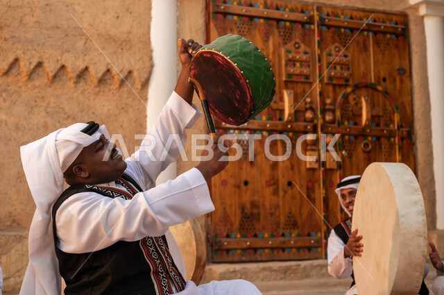 الأحياء التراثية الطينية القديمة، صورة مقربة من الجانب لرجل عربي خليجي سعودي يقوم بالاحتفال بتأسيس الدولة الأولى، الاندماج مع الرقص الشعبي وقرع الطبول احتفالا بيوم بدينا ١٧٢٧، إظهار الولاء والحب للدولة السعودية، ارتداء ازياء العرضه النجدية، عروض الفلكلور الشعبي