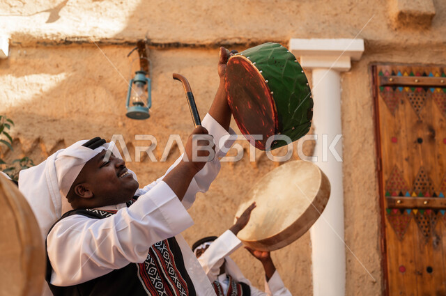 Showing loyalty and love for the Saudi state, ancient mud heritage neighborhoods, a close-up photo from the side of a Saudi Gulf Arab man celebrating the founding of the first state, merging with folk dancing and drumming to celebrate Badina Day 1727, wearing Najdi ardha costumes, folklore performances