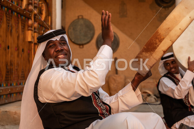 الأحياء التراثية الطينية القديمة، صورة مقربة من الجانب لرجل عربي خليجي سعودي يقوم بالاحتفال بتأسيس الدولة الأولى، الاندماج مع الرقص الشعبي وقرع الطبول احتفالا بيوم بدينا ١٧٢٧، إظهار الولاء والحب للدولة السعودية، ارتداء ازياء العرضه النجدية