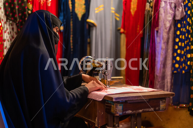 ذكرى تأسيس الدوله السعوديه الاولى ٢٢ فبراير ،يوم التأسيس 1727 م، حرف ومهن تقليدية نسائية، صورة مقربة من الخلف لخياطة عربية سعودية خليجية ترتدي العباءة السوداء والبرقع تقوم بحياكة وخياطة القماش على الماكينة اليدوية في الأسواق الشعبيه القديمة في السعودية