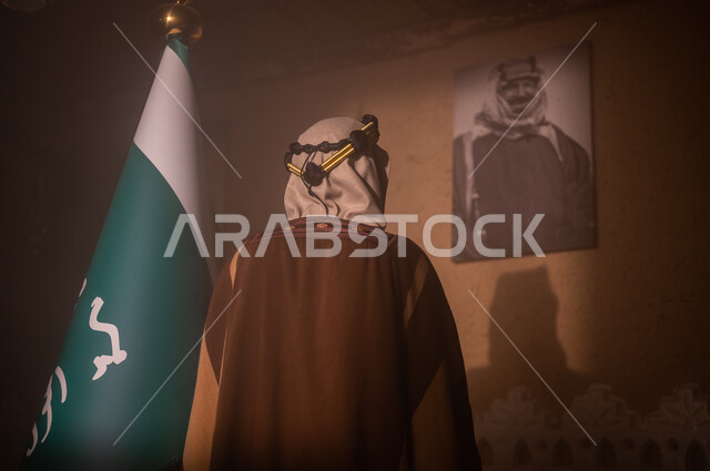 Islam is the banner of monotheism and the emblem of national identity, the concept of pride, pride and love of the homeland, the Day of Our Faith 1727 AD, a close-up photo of a Saudi Gulf Arab man wearing a shemagh and traditional thobe, standing next to the Kingdom’s flag looking at the camera, the anniversary of the founding of the first Saudi state, February 2