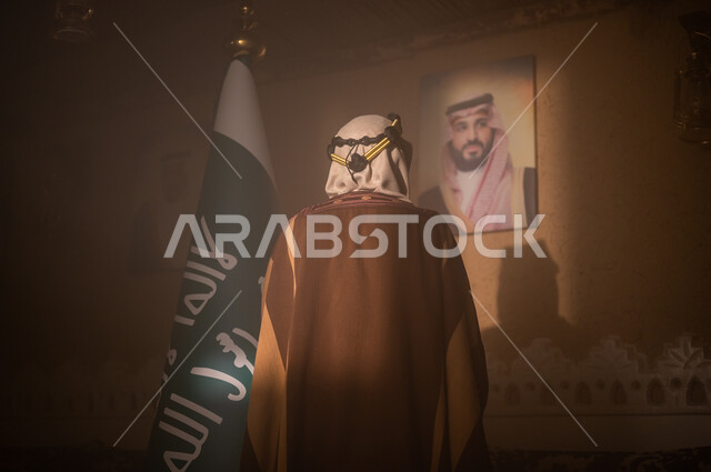 The banner of monotheism and the slogan of national identity, the concept of pride, pride and love of the homeland, the Day of Our Own 1727 AD, a close-up picture from the back of a Saudi Gulf Arab man wearing a shemagh and traditional thobe looking at a picture of Prince Salman bin Abdulaziz standing next to the flag of the Kingdom, the establishment of the first Saudi state, February 22