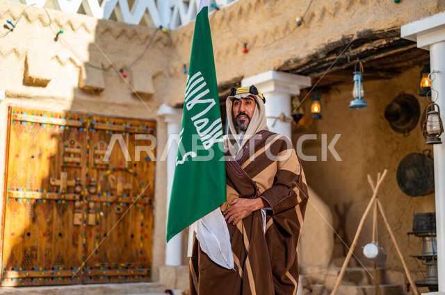 الفخر والاعتزاز بإنجازات الوطن، شعار الهوية الوطنية وراية الإسلام والتوحيد، يوم بدينا 1727 م، رجل عربي خليجي سعودي يرتدي العقال الذهبي والبشت يحمل بيده علم التأسيس في بيت طيني تراثي, ذكرى تأسيس الدولة السعودية الأولى 22 فبراير 
