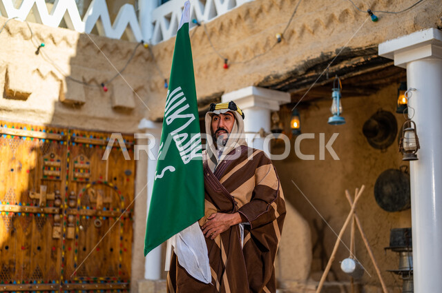 الفخر والاعتزاز بإنجازات الوطن، شعار الهوية الوطنية وراية الإسلام والتوحيد، يوم بدينا 1727 م، رجل عربي خليجي سعودي يرتدي العقال الذهبي والبشت يحمل بيده علم التأسيس في بيت طيني تراثي, ذكرى تأسيس الدولة السعودية الأولى 22 فبراير 