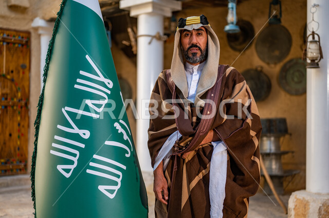 ارتداء الأزياء الشعبية التقليدية، البيوت الطينية التراثية القديمة، شعار الهوية الوطنية وراية الإسلام والتوحيد، يوم بدينا 1727 م، رجل عربي خليجي سعودي يرتدي العقال الذهبي والبشت يحمل بيده علم التأسيس وينظر إلى الكاميرا, ذكرى تأسيس الدولة السعودية الأولى 22 فبراير 