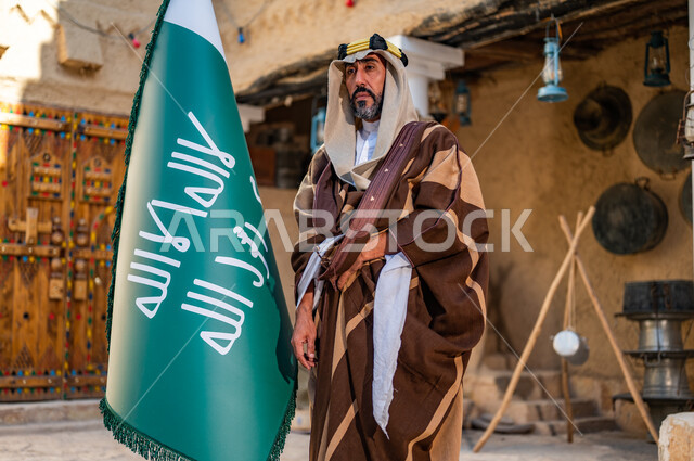 الفخر والاعتزاز بإنجازات الوطن، شعار الهوية الوطنية وراية الإسلام والتوحيد، يوم بدينا 1727 م، رجل عربي خليجي سعودي يرتدي العقال الذهبي والبشت يحمل بيده علم التأسيس في بيت طيني تراثي, ذكرى تأسيس الدولة السعودية الأولى 22 فبراير 