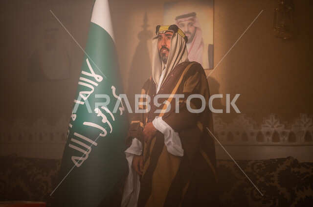 الإسلام  راية التوحيد وشعار الهوية الوطنية, مفهوم الفخر والاعتزاز وحب الوطن، يوم بدينا 1727 م، صورة مقربة لرجل عربي سعودي خليجي يرتدي الشماغ والثوب التقليدي يقف جانب علم المملكة ينظر إلى الكاميرا، ذكرى تأسيس الدولة السعودية الأولى 22 فبراير