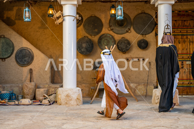 يوم بدينا 1727 م، ارتداء الزي الشعبي في ذكرى تأسيس الدولة السعودية الاولى 22 فبراير، نمط الحياة القديمة، صورة من الخلف لرجل وولد عربيين خليجيين سعوديين يرتديان العقال والبشت التقليدي يتجولان في منزل طيني تراثي