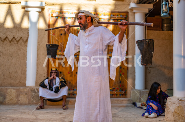  المحافظة على تراث الاجداد و توارثه بين الأجيال، أسلوب الحياة الشعبية البدوية، رجل عربي خليجي سعودي يرتدي الزي التقليدي الشعبي ويحمل على ظهره دلو من الجلد، البيوت الطينية التراثية على الطراز القديم، يوم بدينا 1727 م، ذكرى تأسيس الدولة السعودية الاولى 22 فبراير