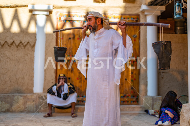  المحافظة على تراث الاجداد و توارثه بين الأجيال، أسلوب الحياة الشعبية البدوية، رجل عربي خليجي سعودي يرتدي الزي التقليدي الشعبي ويحمل على ظهره دلو من الجلد، البيوت الطينية التراثية على الطراز القديم، يوم بدينا 1727 م، ذكرى تأسيس الدولة السعودية الاولى 22 فبراير