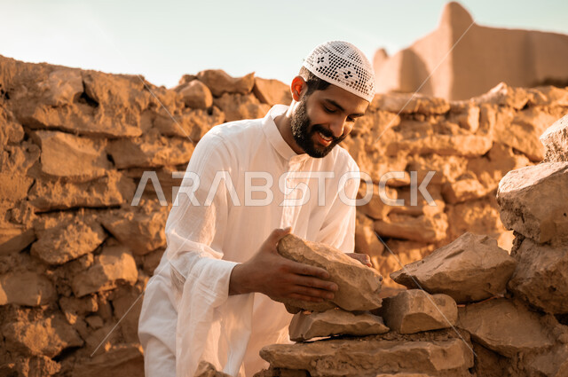 إعادة بناء وترميم البيوت والمنشآت القديمة, مفهوم التمسك بالأصالة والتراث السعودي, شاب عربي خليجي سعودي مبتسم يرتدي الثوب التقليدي والطاقية البيضاء يمسك بيده الحجارة, يوم بدينا 1727م, ذكرى تأسيس الدولة السعودية الأولى في 22 فبراير