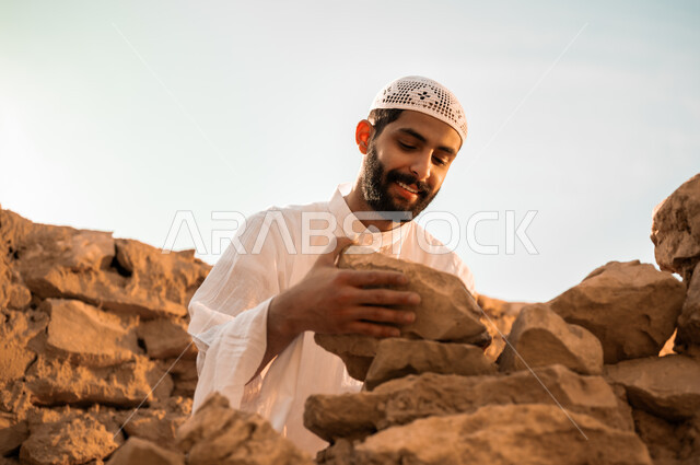 إعادة بناء وترميم البيوت والمنشآت القديمة, مفهوم التمسك بالأصالة والتراث السعودي, شاب عربي خليجي سعودي مبتسم يرتدي الثوب التقليدي والطاقية البيضاء يمسك بيده الحجارة, يوم بدينا 1727م, ذكرى تأسيس الدولة السعودية الأولى في 22 فبراير