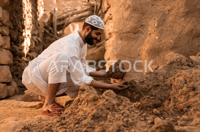 ذكرى تأسيس الدولة السعودية الأولى في 22 فبراير, الفخر والتباهي بالتراث الشعبي السعودي, شاب عربي خليجي سعودي مبتسم يرتدي الطاقية البيضاء والثوب التقليدي يمسك بيده الطين الترميم الحائط, يوم بدينا 1727 م, أحياء وبيوت أثرية قديمة