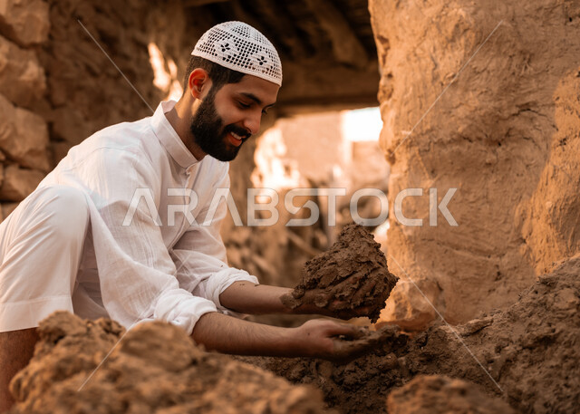 ذكرى تأسيس الدولة السعودية الأولى في 22 فبراير, الفخر والتباهي بالتراث الشعبي السعودي, شاب عربي خليجي سعودي مبتسم يرتدي الطاقية البيضاء والثوب التقليدي يمسك بيده الطين الترميم الحائط, يوم بدينا 1727 م, أحياء وبيوت أثرية قديمة