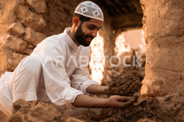 ذكرى تأسيس الدولة السعودية الأولى في 22 فبراير, الفخر والتباهي بالتراث الشعبي السعودي, شاب عربي خليجي سعودي مبتسم يرتدي الطاقية البيضاء والثوب التقليدي يمسك بيده الطين الترميم الحائط, يوم بدينا 1727 م, أحياء وبيوت أثرية قديمة