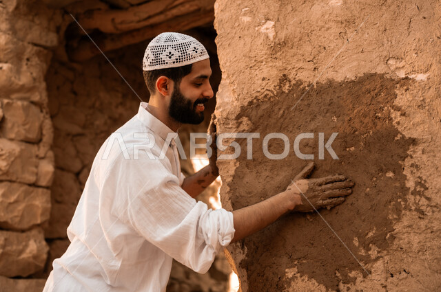 الفخر والتباهي بالتراث الشعبي السعودي, صورة مقربة من الجانب لشاب عربي خليجي سعودي مبتسم يرتدي الطاقية البيضاء والثوب التقليدي يقوم بترميم حائط طيني, يوم بدينا 1727 م, ذكرى تأسيس الدولة السعودية الأولى في 22 فبراير, أحياء وبيوت أثرية قديمة