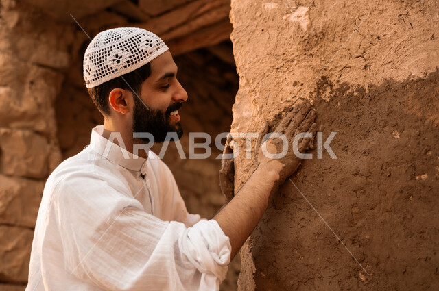 Pride and flaunting of Saudi folklore, a close-up of a smiling Saudi Gulf Arab young man wearing a white hat and traditional dress restoring a mud wall, Day of Dina 1727 AD, anniversary of the founding of the first Saudi state on February 22, ancient neighborhoods and houses