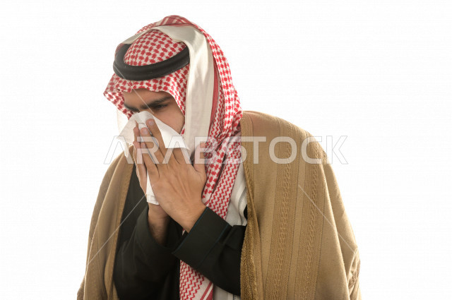 بورتريه لرجل سعودي خليجي يعطس بإيماءات الوجه تدل على المرض، يرتدي ملابس شتوية مناسبة لفصل الشتاء ، الاصابة بالانفلونزا ، خلفية بيضاء