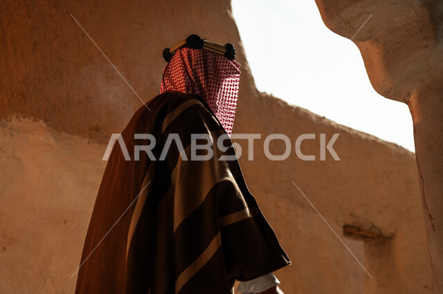 ذكرى تأسيس الدولة السعودية الأولى في 22 فبراير, صورة من الجانب الرجل عربي خليجي سعودي يرتدي البشت والعقال والشماغ يقف في إحدى الأحياء والمنشآت الطينية التراثية القديمة, ارتداء الأزياء الشعبية التقليدية في الأعياد والمناسبات الوطنية, يوم بدينا 1727 م