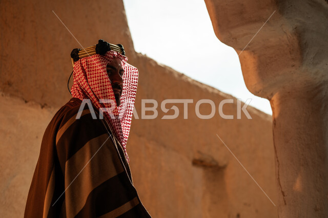 ذكرى تأسيس الدولة السعودية الأولى في 22 فبراير, صورة من الجانب الرجل عربي خليجي سعودي يرتدي البشت والعقال والشماغ يقف في إحدى الأحياء والمنشآت الطينية التراثية القديمة, ارتداء الأزياء الشعبية التقليدية في الأعياد والمناسبات الوطنية, يوم بدينا 1727 م