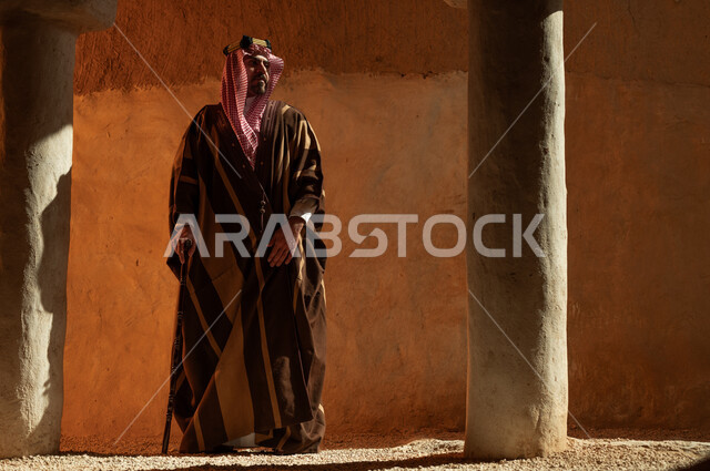 الوقوف بالأحياء والمنشآت الطينية التراثية القديمة, صورة من الجانب الرجل عربي خليجي سعودي يرتدي البشت والعقال يحمل العكاز, ارتداء الأزياء الشعبية التقليدية في الأعياد والمناسبات الوطنية, يوم بدينا 1727 م, ذكرى تأسيس الدولة السعودية الأولى في 22 فبراير