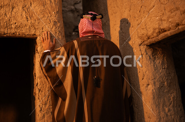 ذكرى تأسيس الدولة السعودية الأولى في 22 فبراير, الفخر بالأصالة والتراث السعودي, صورة مقربة من الخلف لرجل عربي خليجي سعودي يرتدي البشت والعقال يقف في إحدى البيوت والأحياء الطينية القديمة, ارتداء الأزياء الشعبية في الأعياد والمناسبات الوطنية, يوم بدينا 1727م