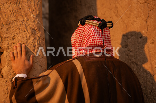 يوم بدينا 1727م, ذكرى تأسيس الدولة السعودية الأولى في 22 فبراير, الفخر بالأصالة والتراث السعودي, صورة مقربة من الخلف لرجل عربي خليجي سعودي يرتدي البشت والعقال يقف في إحدى البيوت والأحياء الطينية القديمة, ارتداء الأزياء الشعبية في الأعياد والمناسبات الوطنية