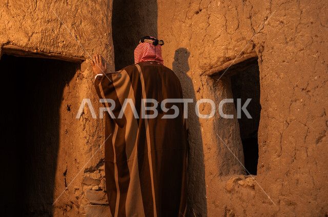 الاعتزاز والشموخ والفخر بالتراث السعودي, صورة من الخلف لرجل عربي خليجي سعودي يرتدي البشت والعقال يقف في إحدى البيوت والأحياء الطينية القديمة, ارتداء الأزياء الشعبية في الأعياد والمناسبات الوطنية, يوم بدينا 1727م, ذكرى تأسيس الدولة السعودية الأولى في 22 فبراير