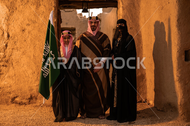 رفع علم المملكة بفخر، ذكرى تأسيس الدولة السعودية الاولى 22 فبراير، عائلة عربيه خليجية سعودية ترتدي الأزياء الشعبية في الأعياد والمناسبات الوطنية يقفون في إحدى البيوت التراثية المبنية من الطين، النظر إلى الكاميرا بإيماءات الاعتزاز والشموخ، يوم بدينا 1727 م