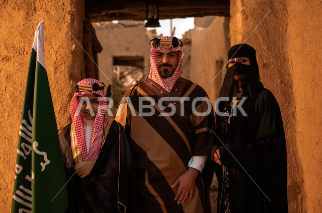 رفع علم المملكة بفخر، ذكرى تأسيس الدولة السعودية الاولى 22 فبراير، عائلة عربيه خليجية سعودية ترتدي الأزياء الشعبية في الأعياد والمناسبات الوطنية يقفون في إحدى البيوت التراثية المبنية من الطين، النظر إلى الكاميرا بإيماءات الاعتزاز والشموخ، يوم بدينا 1727 م