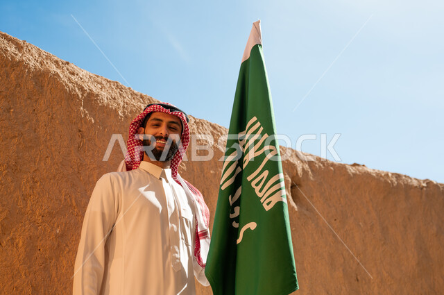 تأسيس الدولة السعودية الأولى في 22 فبراير, مشاعر الفخر والانتماء للوطن, الاحياء والبيوت التراثية القديمة, يوم بدينا 1727م, صورة مقربة لرجل عربي خليجي سعودي يرتدي الثوب التقليدي والشماغ يحمل علم التأسيس باعتزاز