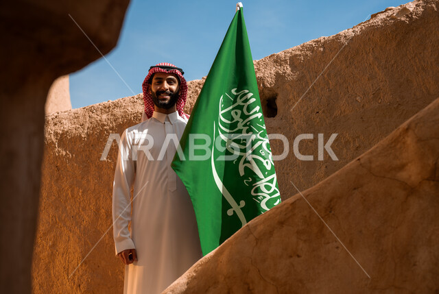 يوم العلم 11 مارس, ذكرى تأسيس الدولة السعودية الأولى في 22 فبراير, اظهار مشاعر الفخر والانتماء للوطن, الاحياء والبيوت التراثية القديمة, يوم بدينا 1727م, رجل عربي خليجي سعودي يرتدي الثوب التقليدي والشماغ يحمل علم المملكة باعتزاز في العيد الوطني في 23 سبتمبر