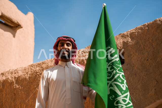 تأسيس الدولة السعودية الأولى في 22 فبراير, مشاعر الفخر والانتماء للوطن, الاحياء والبيوت التراثية القديمة, يوم بدينا 1727م, صورة مقربة لرجل عربي خليجي سعودي يرتدي الثوب التقليدي والشماغ يحمل علم المملكة باعتزاز في العيد الوطني في 23 سبتمبر, يوم العلم 11 مارس