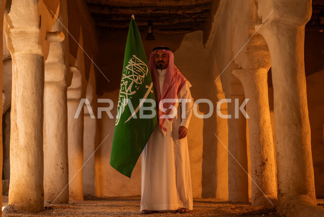 Showing feelings of pride and pride in national identity on the anniversary of the founding of the first Saudi state on February 22, old heritage installations and columns, Day of Dina 1727 AD, a Saudi Gulf Arab man wearing the traditional thobe and shemagh raising the flag on the Saudi National Day September 23, Flag Day March 11