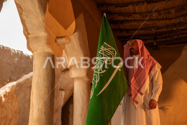 اظهار مشاعر الفخر والاعتزاز بالهوية الوطنية في ذكرى تأسيس الدولة السعودية الأولى في 22 فبراير, المنشآت والأعمدة التراثية القديمة, يوم بدينا 1727م, رجل عربي خليجي سعودي يرتدي الثوب التقليدي والشماغ يرفع العلم في العيد الوطني السعودي 23 سبتمبر, يوم العلم 11 مارس