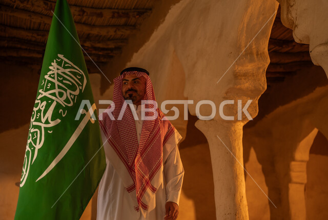 المنشآت والأعمدة التراثية القديمة, اظهار مشاعر الفخر والاعتزاز بالهوية الوطنية في ذكرى تأسيس الدولة السعودية الأولى في 22 فبراير, يوم بدينا 1727م, رجل عربي خليجي سعودي يرتدي الثوب التقليدي والشماغ يرفع العلم في العيد الوطني السعودي 23 سبتمبر, يوم العلم 11 مارس