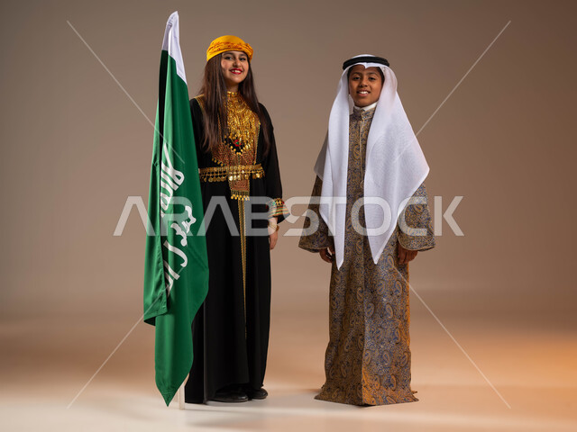يوم بدينا 1727م, ذكرى تأسيس الدولة السعودية الأولى في 22 فبراير, تعليم الأولاد الاعتزاز بالانتماء للوطن، بورتريه لولد وفتاة عرب خليجيين سعوديين يرتديان أزياء المنطقة الوسطة والجنوبية, الوقوف بجانب علم التأسيس بفخر, صورة الجسم بالكامل, خلفية بيج