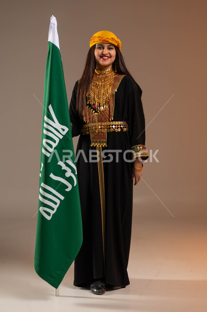 ذكرى تأسيس الدولة السعودية الأولى في 22 فبراير, ارتداء أزياء المنطقة الجنوبية في المناسبات الوطنية, بورتريه لفتاة عربية خليجية سعودية ترتدي الشيلة والسيدرة تنظر إلى الكاميرا وتمسك علم الأسيس بفخر, يوم بدينا 1727م, صورة الجسم بالطول الكامل, خلفية بيج