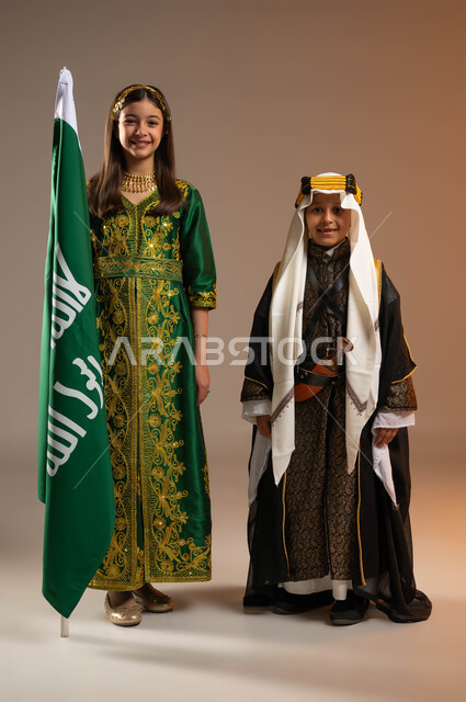 يوم بدينا 1727م, ذكرى تأسيس الدولة السعودية الأولى في 22 فبراير, الوقوف باحترام بجانب علم يوم التأسيس, إحياء التراث الخليجي, بورتريه لولد وفتاة عربيين خليجيين سعوديين يرتديان الزي الشعبي ينظران إلى الكاميرا, صورة الجسم بالكامل, خلفية بيج
