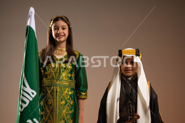 الوقوف باحترام بجانب علم يوم التأسيس, إحياء التراث الخليجي, بورتريه مقرب لولد وفتاة عربيين خليجيين سعوديين يرتديان الزي الشعبي ينظران إلى الكاميرا, التباهي والفخر بالانتماء للوطن, يوم بدينا 1727م, ذكرى تأسيس الدولة السعودية الأولى في 22 فبراير, خلفية بيج
