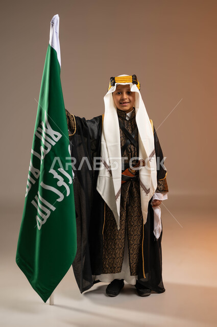 الوقوف بفخر بإنجازات البلاد, النظر إلى الكاميرا بتباهي واعتزاز, بورتريه لولد عربي خليجي سعودي يرتدي البشت والعقال والدقلة يمسك علم يوم التأسيس بتعابير السرور, يوم بدينا 1727م, ذكرى تأسيس الدولة السعودية الأولى في 22 فبراير, صورة الجسم بالطول الكامل, خلفية بيج
