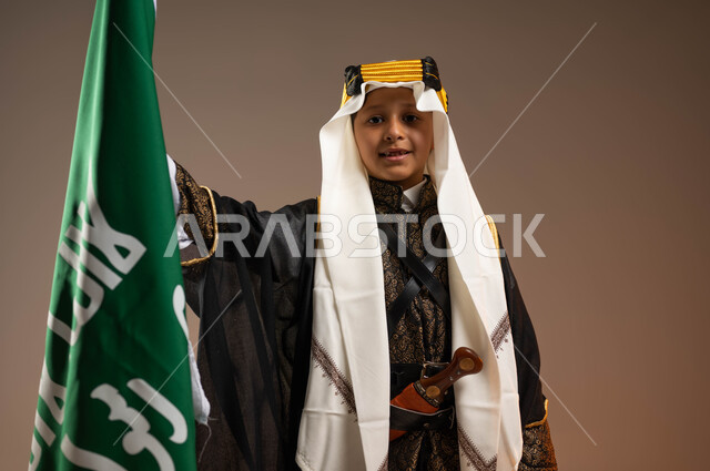 الوقوف بفخر بإنجازات البلاد, النظر إلى الكاميرا بتباهي واعتزاز, بورتريه مقر ب لولد عربي خليجي سعودي يرتدي البشت والعقال والدقلة يمسك علم يوم التأسيس بتعابير السرور, يوم بدينا 1727م, ذكرى تأسيس الدولة السعودية الأولى في 22 فبراير, خلفية بيج