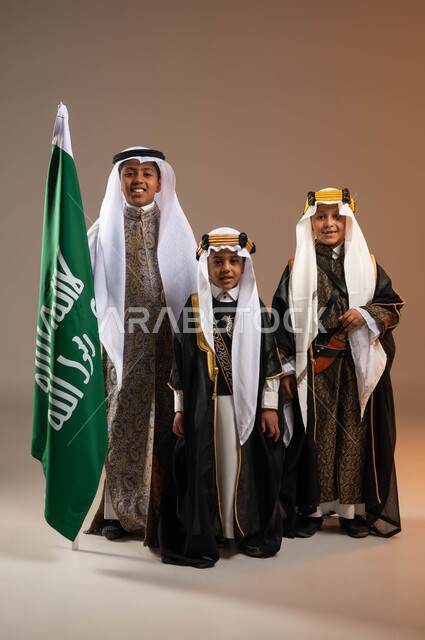 الوقوف بفخر بجانب علم التأسيس, يوم بدينا 1727 م، النظر إلى الكاميرا بتعابير الفرح والسرور، بورتريه لمجموعة أولاد عرب خليجيين سعوديين يرتدون الأزياء الشعبية التقليدية، ذكرى تأسيس الدولة السعودية 22 فبراير، صورة الجسم بالطول الكامل, خلفية بيج