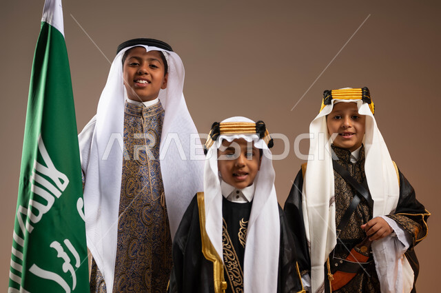 الوقوف بفخر بجانب علم التأسيس, تعليم الأبناء الاعتزاز بأنجازات الوطن، يوم بدينا 1727 م، النظر إلى الكاميرا بتعابير الفرح والسرور، بورتريه مقرب لمجموعة أولاد عرب خليجيين سعوديين يرتدون الأزياء الشعبية التقليدية، ذكرى تأسيس الدولة السعودية 22 فبراير، خلفية بيج