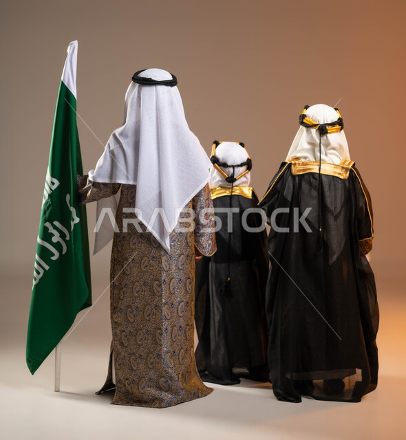 Children participating in national occasions, a portrait from the back of a group of Gulf Arab Saudi boys wearing traditional folk costumes, standing next to the founding flag with pride and honor, Day of Our Day 1727 AD, the anniversary of the founding of the first Saudi state on February 22, full-length body portrait, beige background