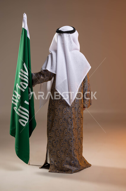 ذكرى تأسيس الدولة السعودية الأولى في 22 فبرير, ارتداء أزياء المنطقة الوسطى في المناسبات الوطنية, بورتريه من الخلف لولد عربي خليجي سعودي يرتدي الدقلة والعقال الشعبي يقف بجانب علم التأسيس, يوم بدينا 1727م, صورة الجسم بالكامل, خلفية بيج