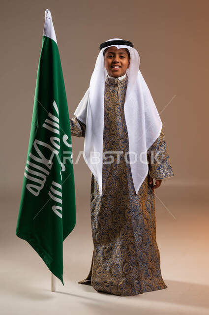 ذكرى تأسيس الدولة السعودية 22 فبراير، بورتريه لولد عربي خليجي سعودي مبتسم يرتدي الدقلة والعقال التقليدي الشعبي يقف بجانب علم التأسيس بإيماءات السعادة، يوم بدينا 1727م، ارتداء أزياء المنطقة الوسطى في المناسبات الوطنية, صورة الجسم بالكامل, خلفية بيج